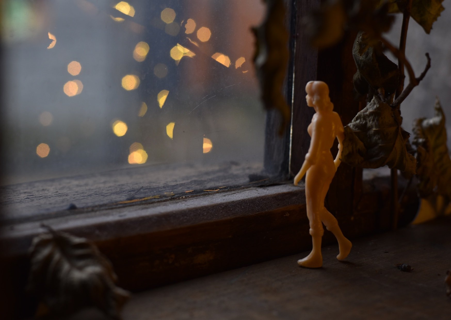 Perfectly strange... open call image, featuring a pale orange plastic miniature figure gazing out of a dark window towards a starlit evening.